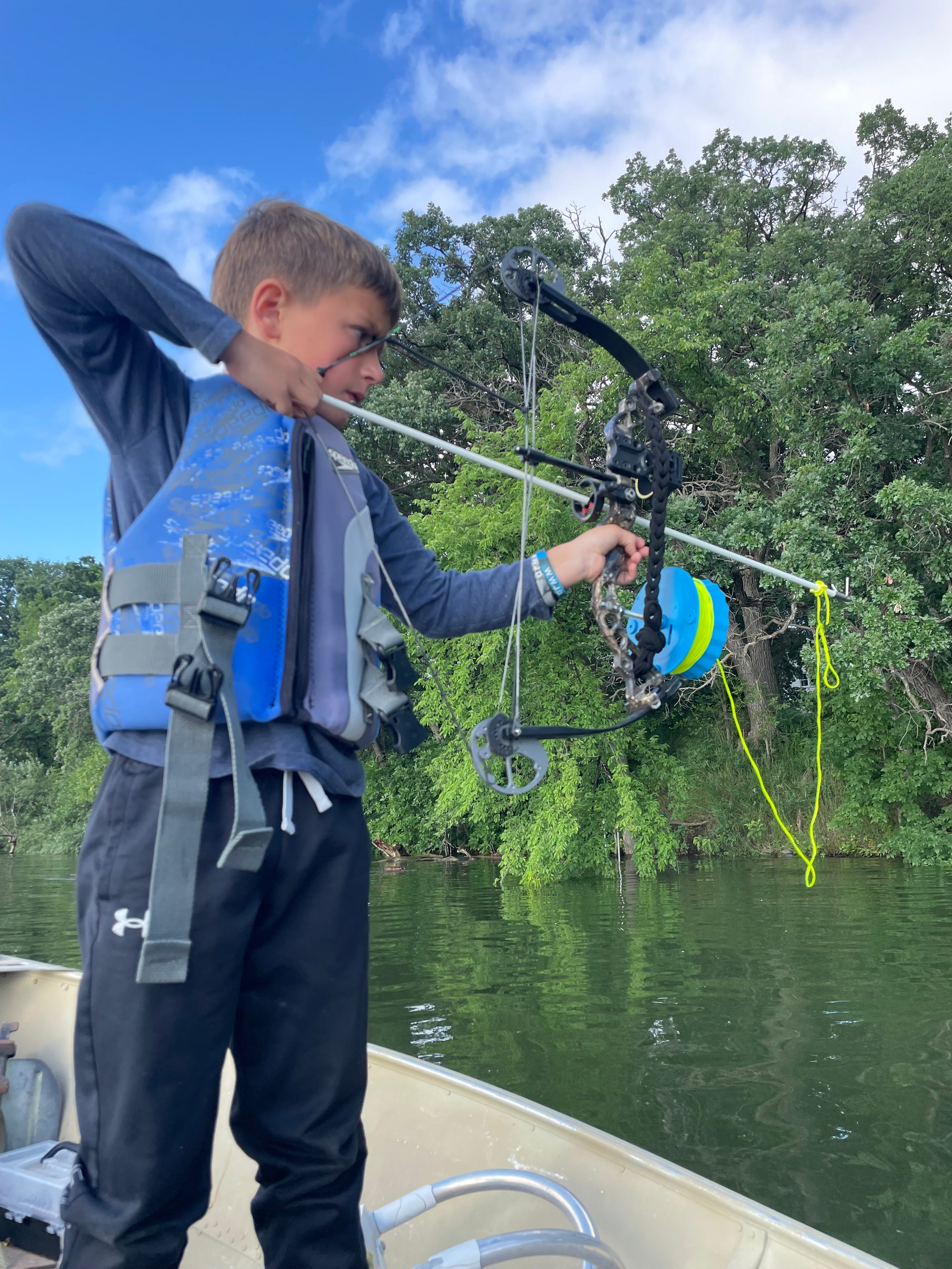 Person preparing to shoot a bow and arrow at fish with Vsling outdoors with trees and sky in the background