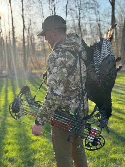 Man in camouflage holding a bow and a dead turkey in a forest setting with black and green Vsling