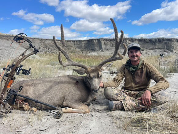 Man posing with a large deer he has hunted, with a bow and his Vsling in the background.